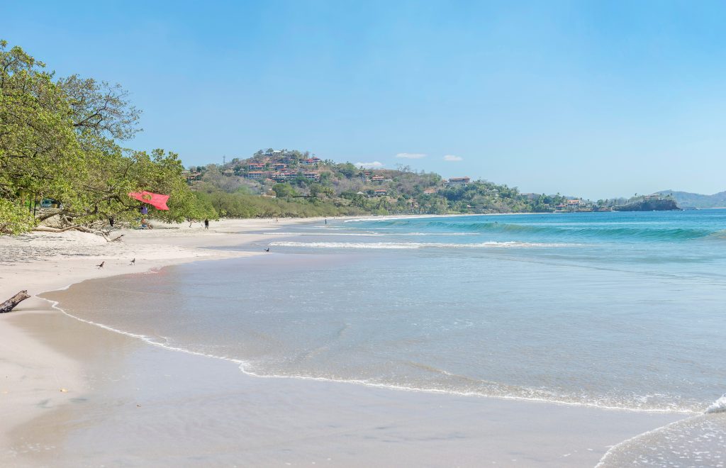 Sandy shoreline at Playa Flamingo Costa Rica with calm waves and hillside homes reflecting the Playa Flamingo lifestyle