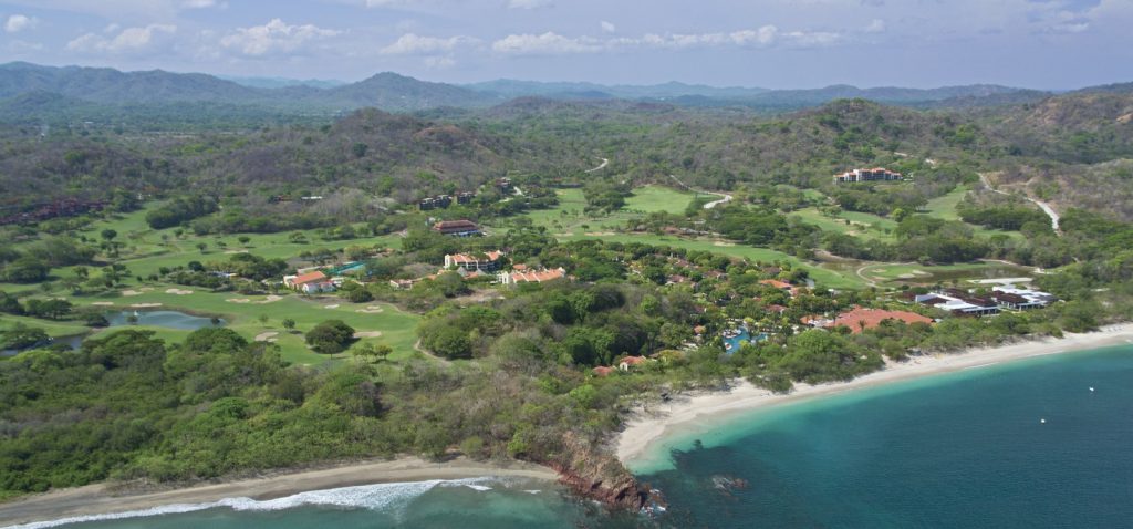 Aerial view of the Reserva Conchal golf course and Playa Conchal coastline surrounded by lush hills.