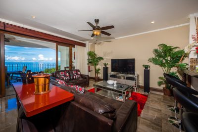 Photograph of a spacious living room in Penthouse 32 with leather sofas, a TV, and a bar area, with large sliding glass doors opening to a balcony with a panoramic ocean view.