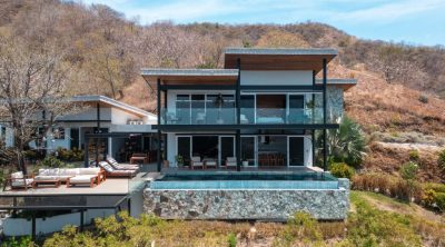 Modern two-level home with glass railings, infinity-edge pool, and outdoor seating set against a hillside in Las Ventanas, Playa Grande