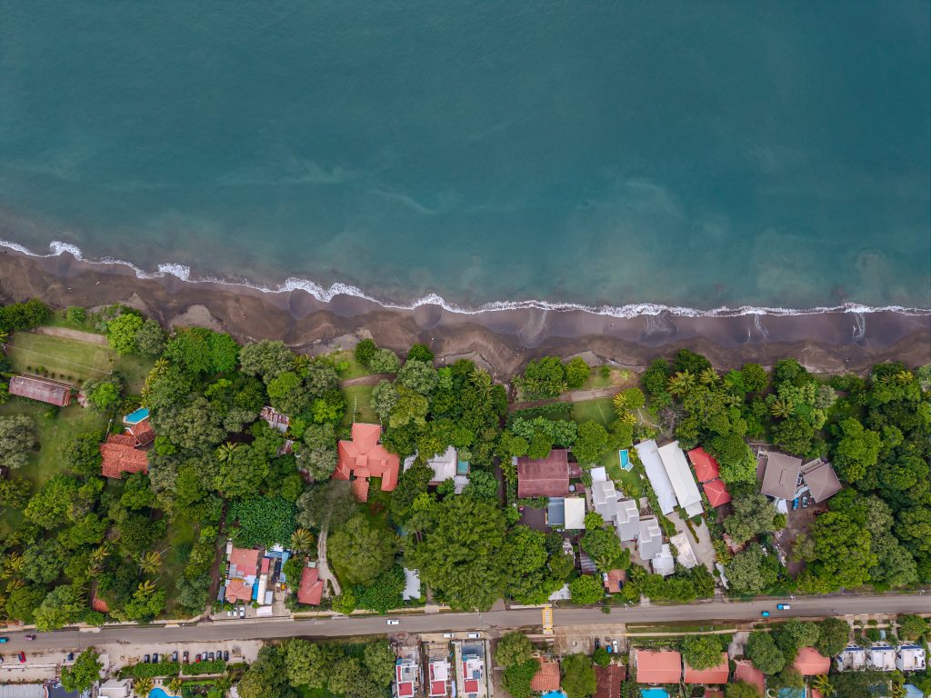 Aerial view of homes along the shoreline in Playas del Coco, with tree-lined streets, dark sand beach, and calm ocean water.