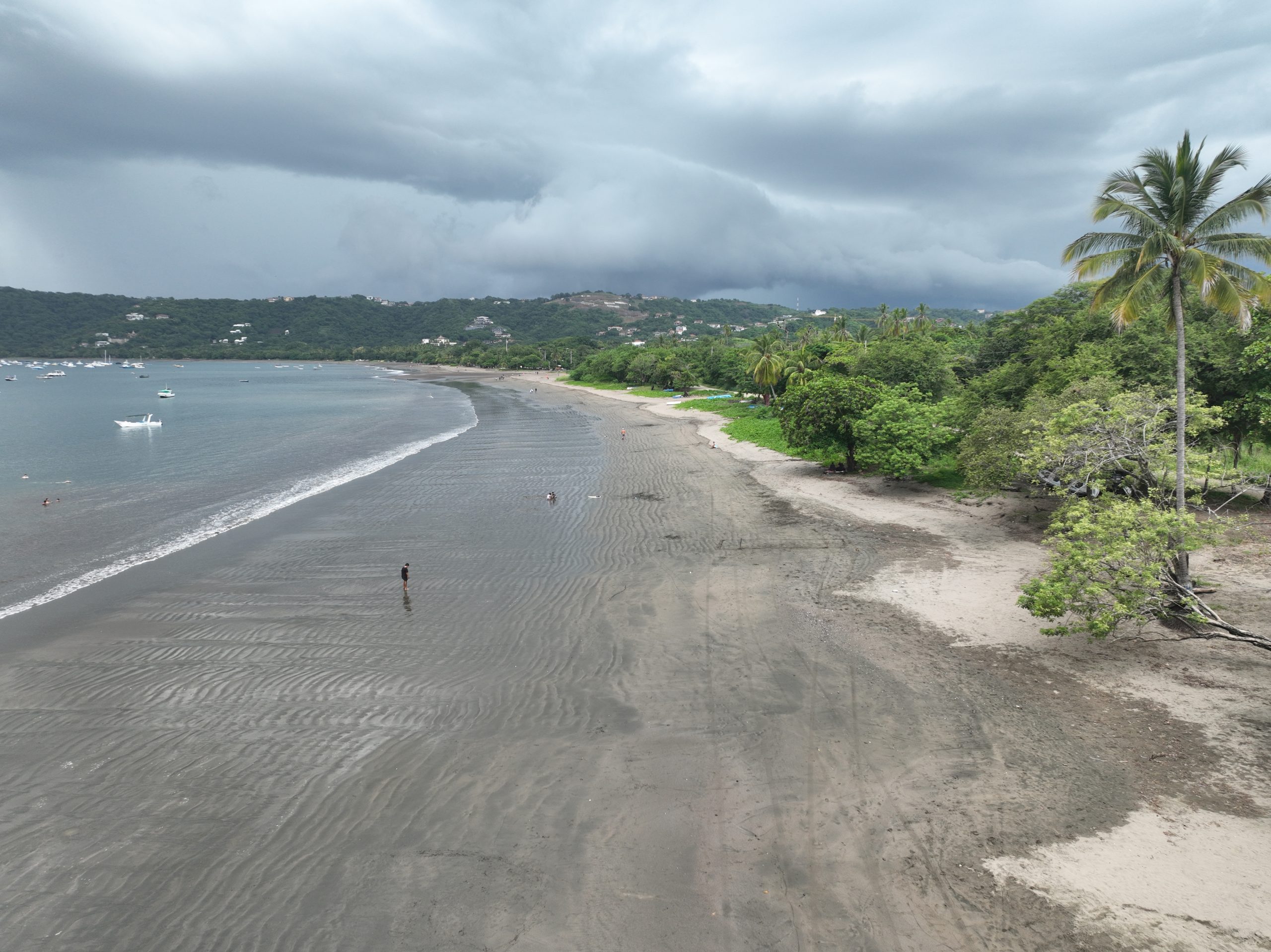 Wide beach view in Playas del Coco with calm shoreline, anchored boats, and green hills in the background under cloudy skies.