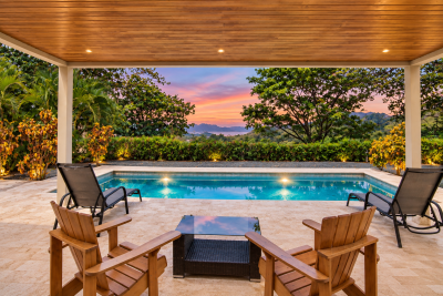 Covered terrace with wooden chairs facing a blue pool, tropical trees, distant hills, and a pink sunset.