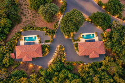 Top-down aerial view of two red-roof homes, each with a rectangular private pool, surrounded by tropical trees.