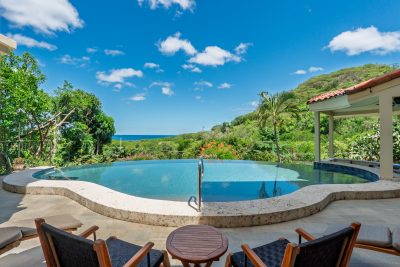 Freeform infinity pool on a tiled patio with wooden lounge chairs overlooking the ocean horizon under a clear blue sky.