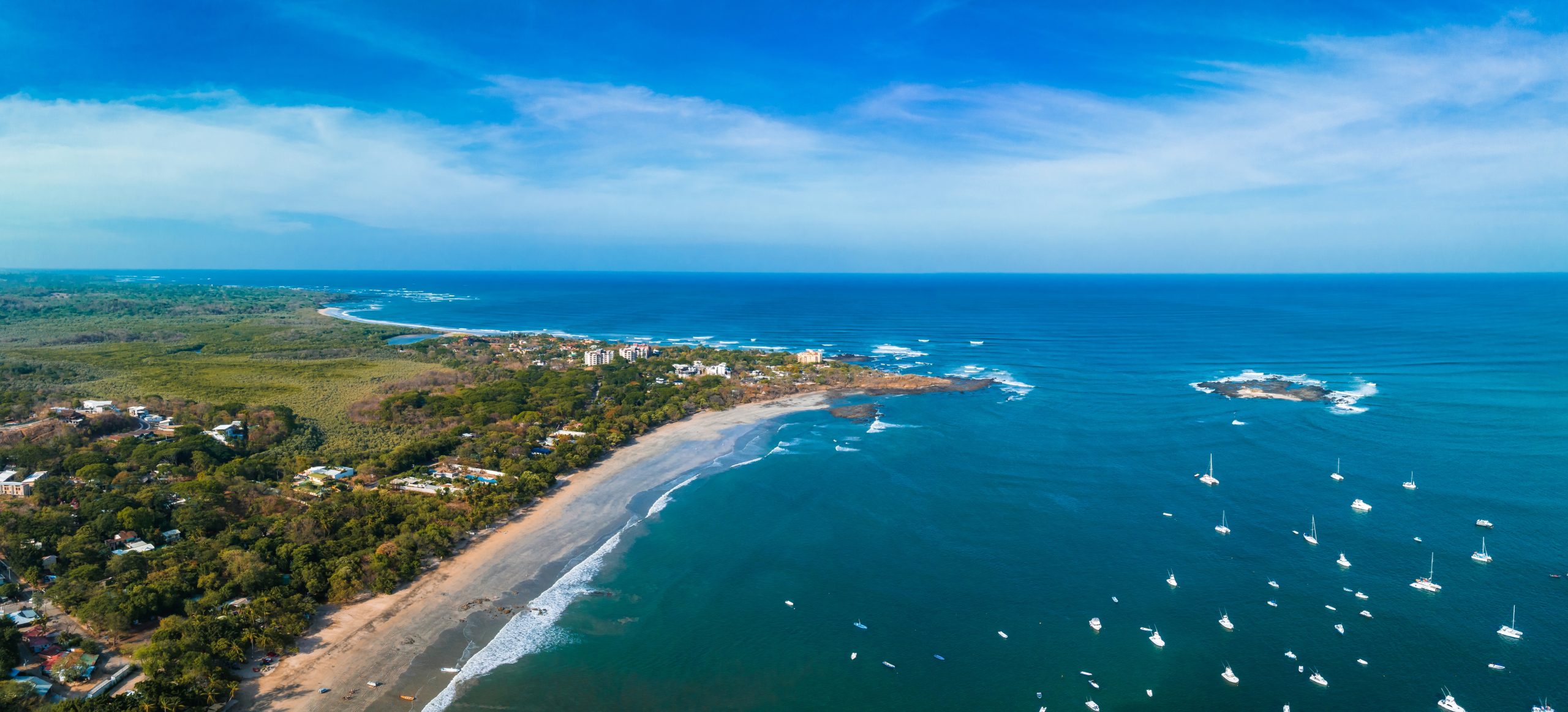 Aerial view of Playa Potrero’s coastline, sandy beach, forested hills, blue ocean, and anchored boats in Guanacaste.