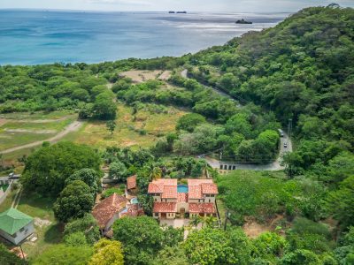 Aerial view of a hillside property surrounded by green forest overlooking the blue waters of Playa Potrero and the Pacific Ocean.
