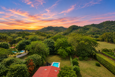 Aerial view of Casa Girasol’s red roof, swimming pool, green grounds, tropical trees, and hills at sunset.