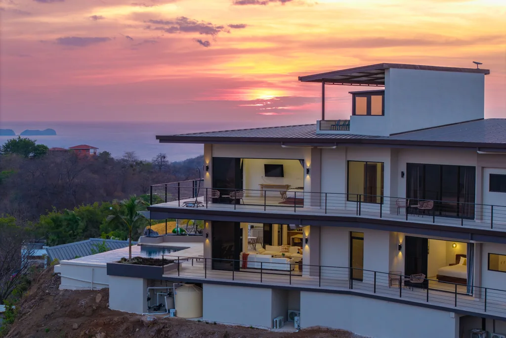Modern multi-level ocean-view home at sunset with lit interiors, infinity pool, and terraces overlooking the Pacific in Guanacaste, Costa Rica
