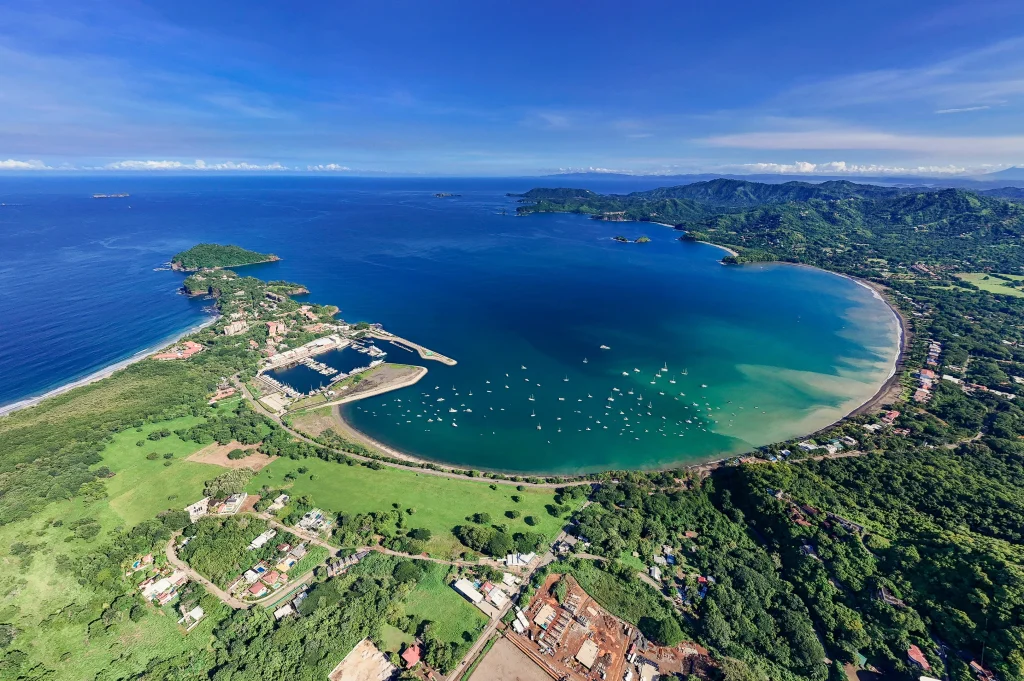An aerial view highlights Playa Flamingo's coastline, white-sand beach, marina, surrounding hills, and Pacific Ocean setting.