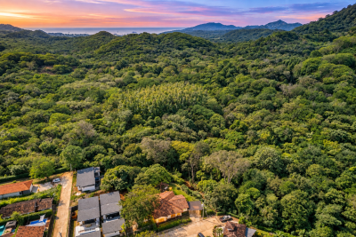 Aerial view of Casa Monos beside dense tropical forest with surrounding homes, hills, and the Pacific coast at sunset.