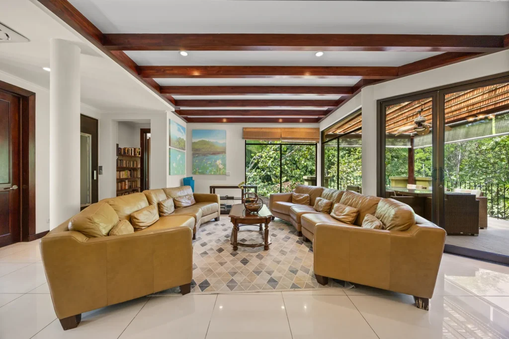 Open-plan living room with wood-beam ceiling opening onto tropical forest through floor-to-ceiling glass, in a Reserva Conchal home