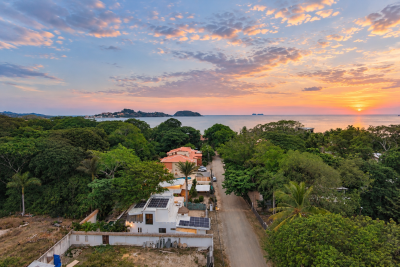 Aerial view over Surfside toward the Pacific Ocean at sunset, with tropical greenery, homes, and a residential road.